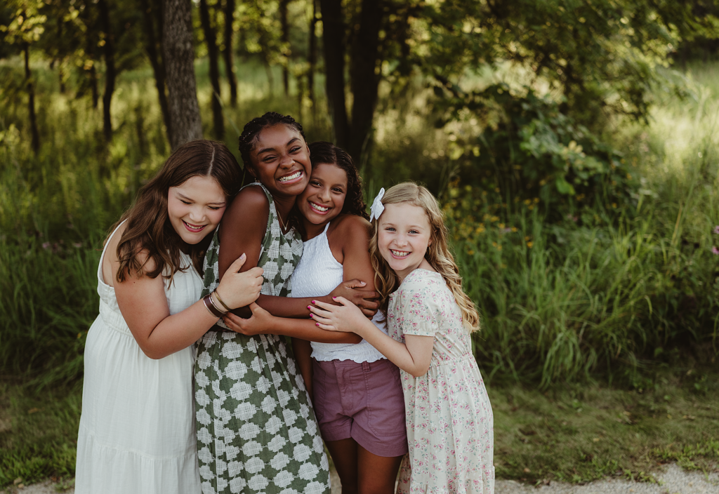 Four smiling girls hugging each other during a joyful outdoor family photography session at a wooded park in the Chicagoland area, captured by LibbyLou Images.