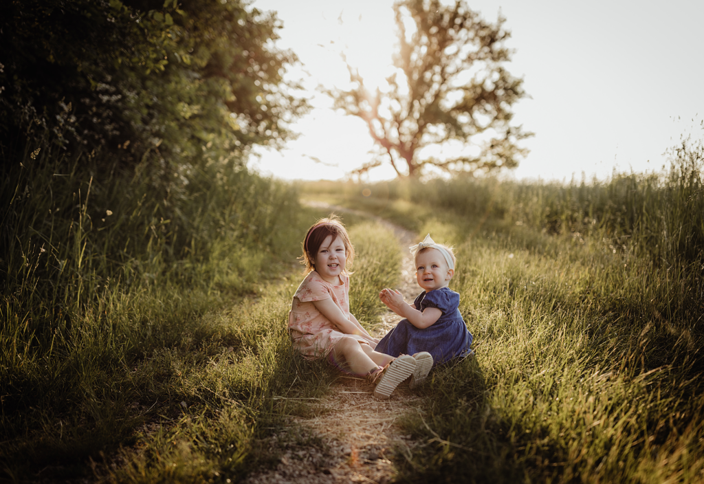 Two young sisters sitting on a grassy trail during a golden hour family photography session at Deer Grove East Forest Preserve in Palatine, Illinois.