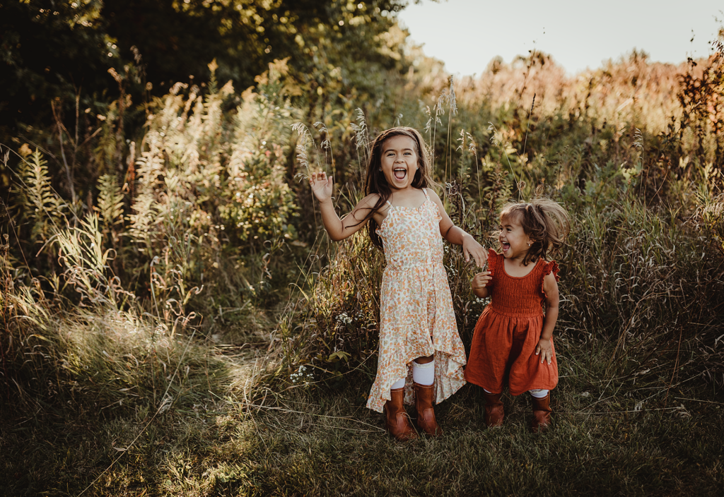 Two young sisters laughing together in a grassy field during a family photo session at Burnidge Forest Preserve in Elgin, Illinois.