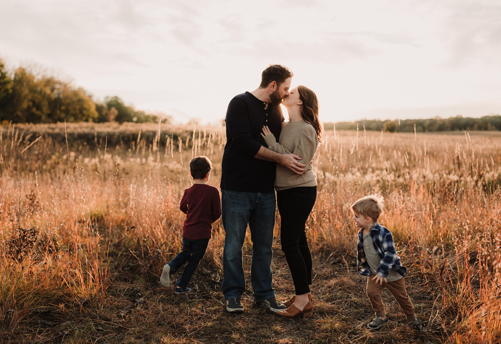 Parents sharing a kiss while their children play nearby during a family session at Deer Grove East in Palatine, Illinois.