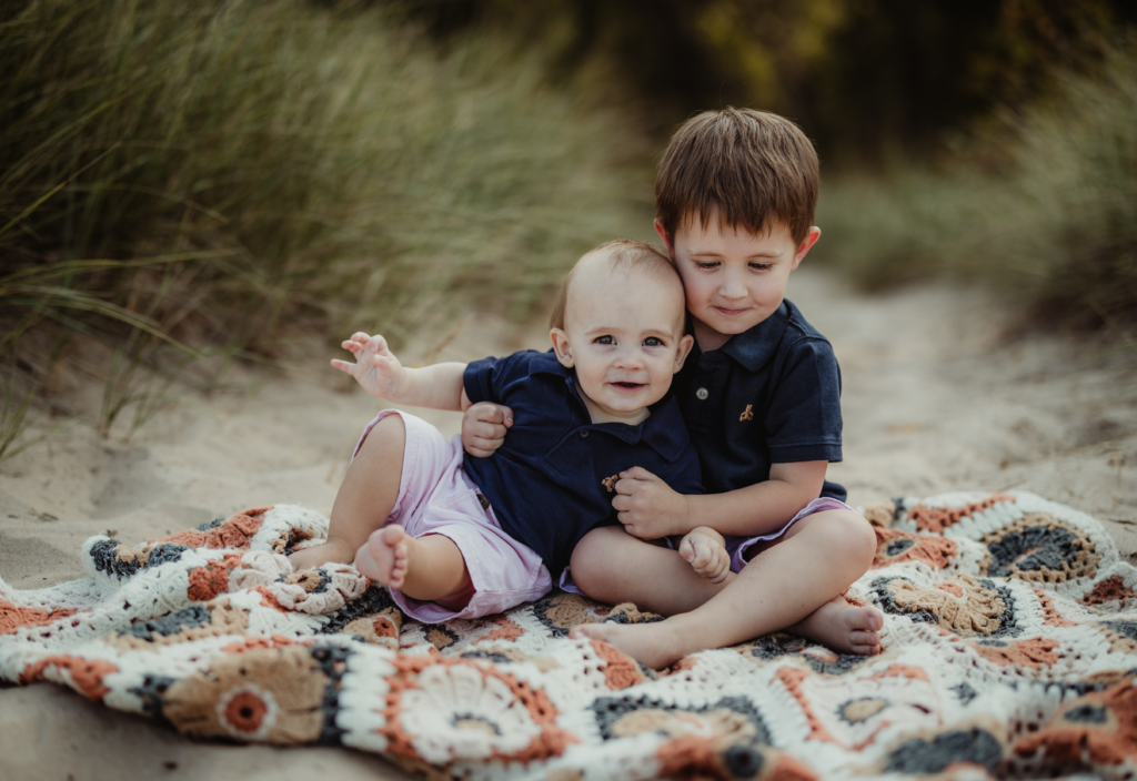 Older brother hugging his baby sibling while sitting on a blanket at Lighthouse Beach in Evanston, IL.