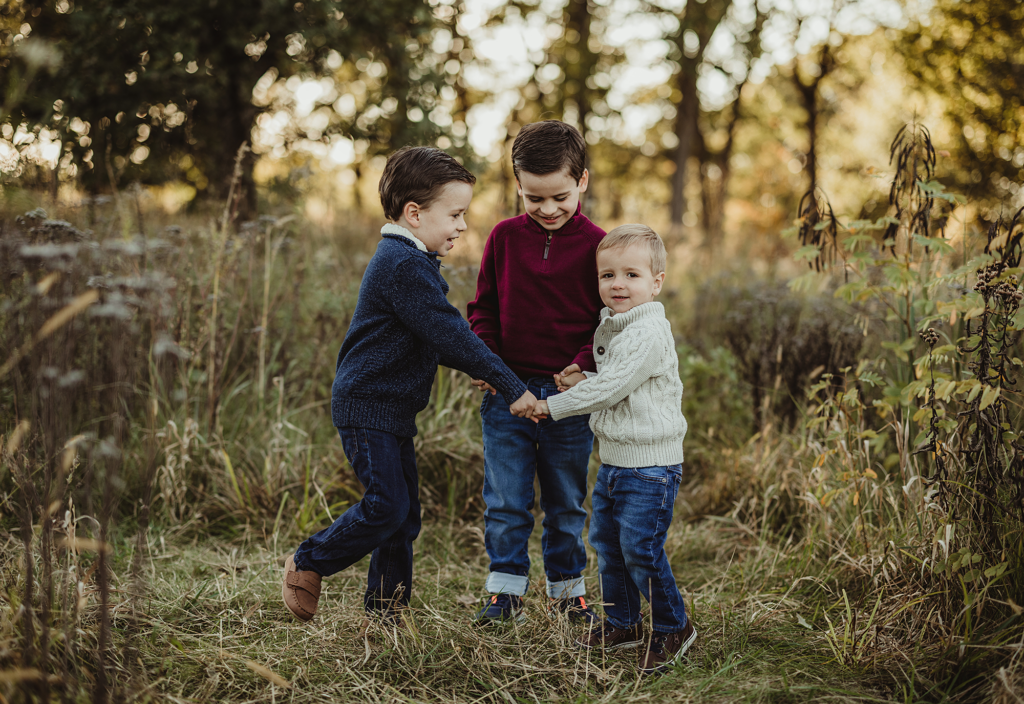 Three young brothers holding hands and playing together during a family session at Mayslake Forest Preserve in Oak Brook, Illinois.