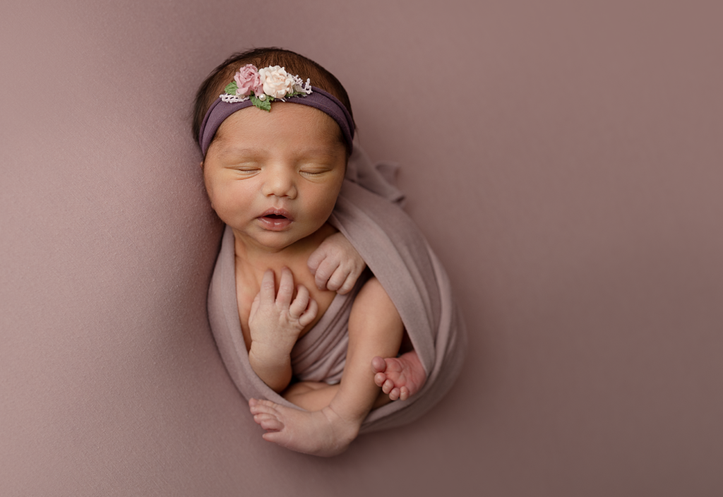 Newborn baby girl wrapped in mauve with a floral headband during her studio session