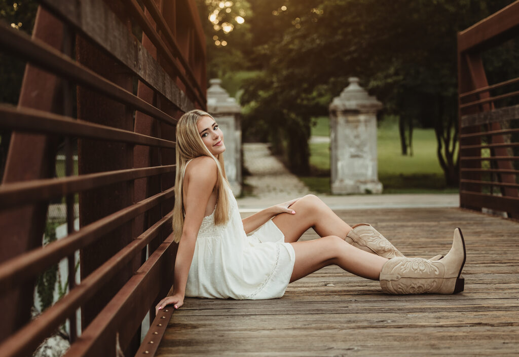 Central High School senior in a white dress and cowboy boots sitting on the bridge at Fabyan Forest Preserve