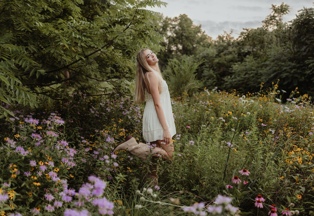 Senior girl in a white dress and cowboy boots surrounded by wildflowers at Fabyan Forest Preserve in Geneva, IL