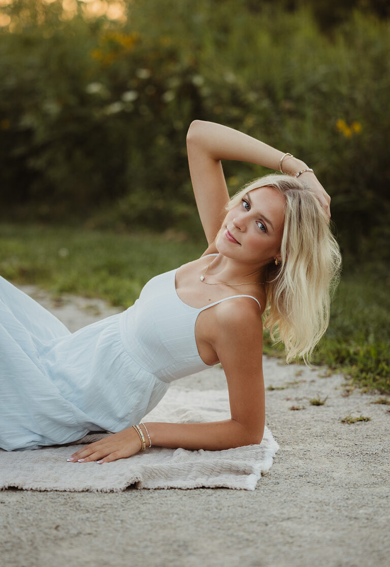 High school senior in a light blue dress reclining on a blanket beside wildflowers at golden hour