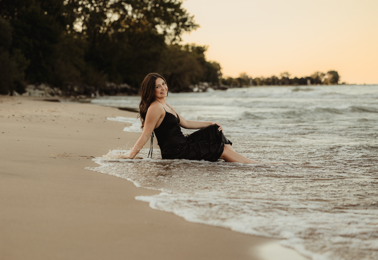Central High school senior in a black dress sitting along the shoreline at Lighthouse Beach in Evanston, Illinois