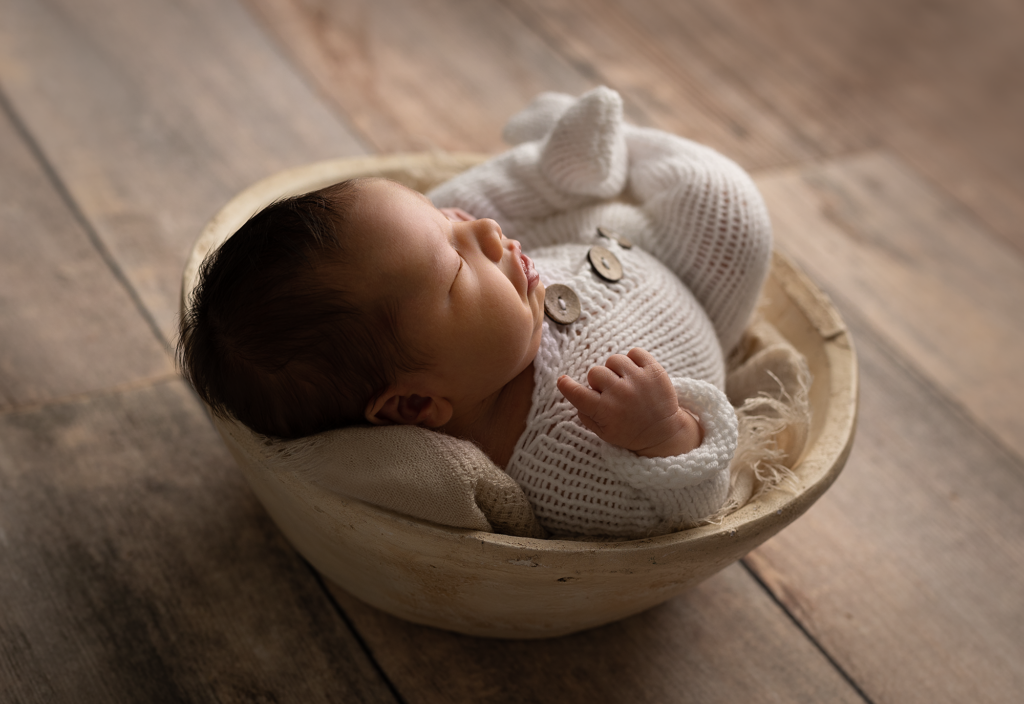 Sleeping newborn baby boy wearing a white knit outfit while posed in a wooden bowl