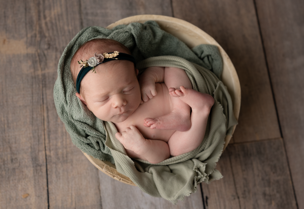 Sleeping newborn baby girl posed in sage green inside a wooden bowl