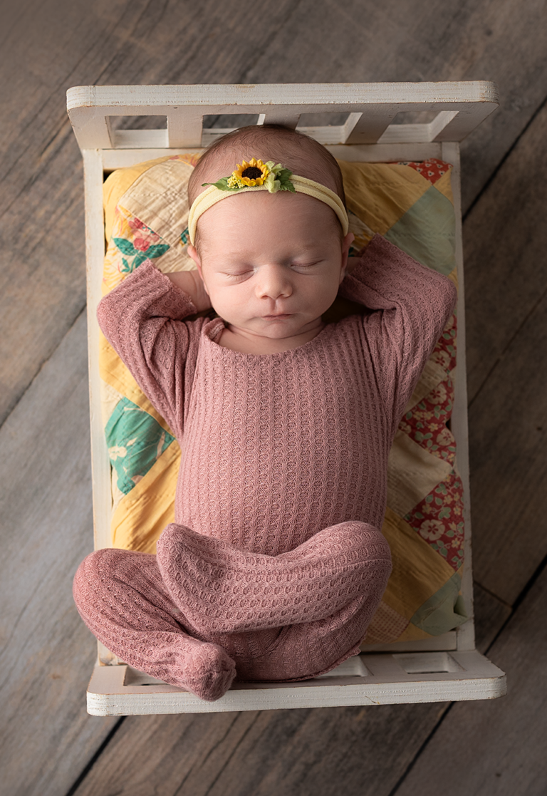 Newborn baby girl wearing a dusty rose outfit while sleeping in a miniature vintage bed