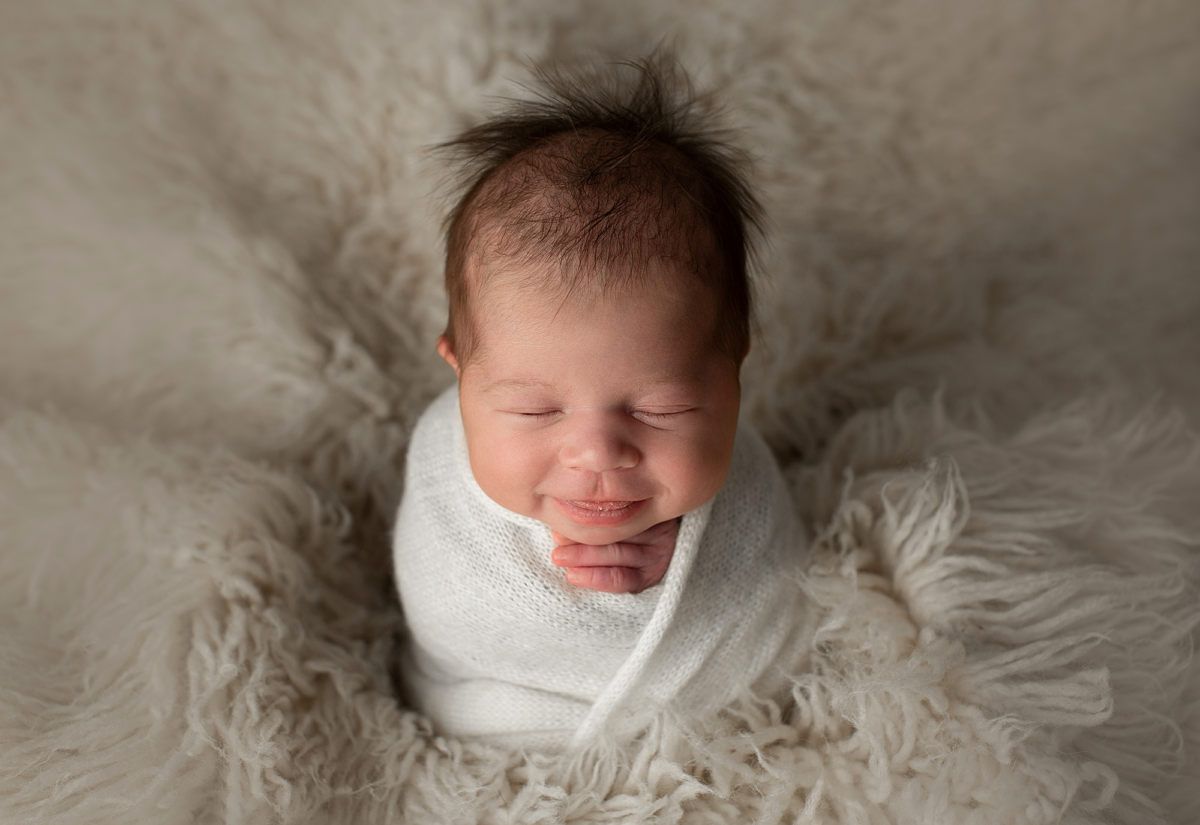 Smiling newborn baby wrapped in white and surrounded by soft cream textures
