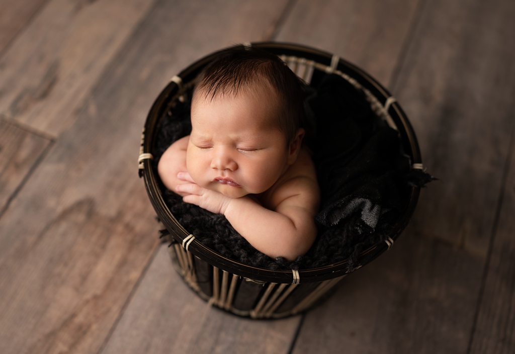 Sleeping newborn baby boy posed with his hands under his chin in a black basket