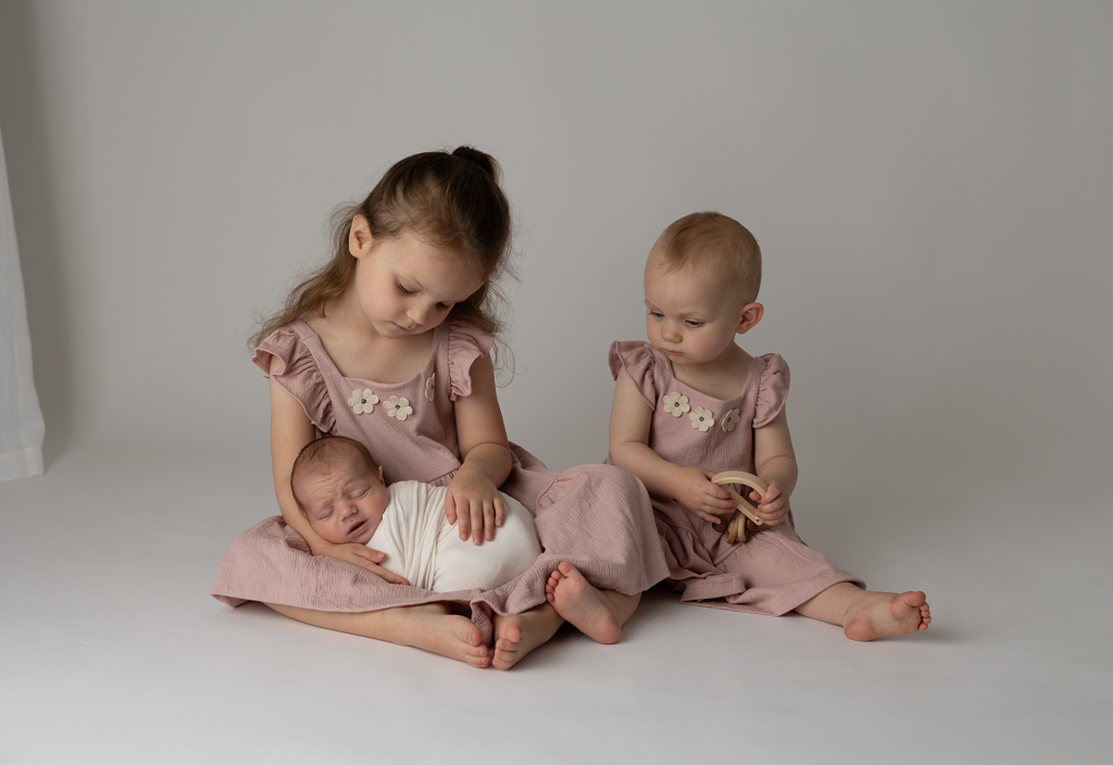 Two older sisters sitting with their swaddled newborn sibling during an Elgin studio newborn session
