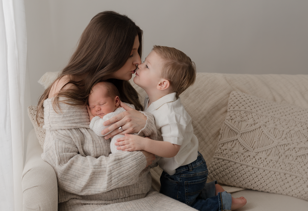 Mother holding her newborn while big brother gives her a kiss during a newborn session