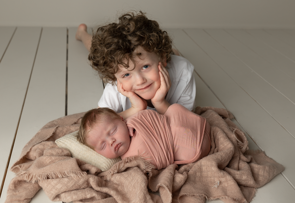 Older brother smiling beside his newborn sibling wrapped in soft pink during a newborn portrait session