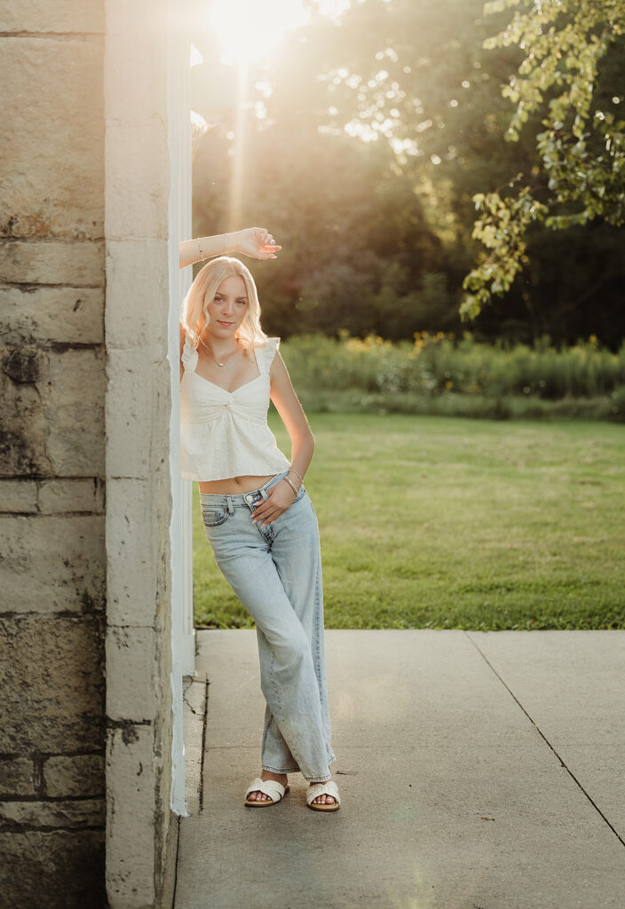 Blonde high school senior leaning against a stone building with warm evening sunlight behind her