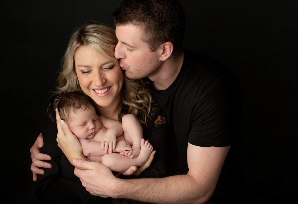 Parents cuddling their newborn together during a black background newborn portrait