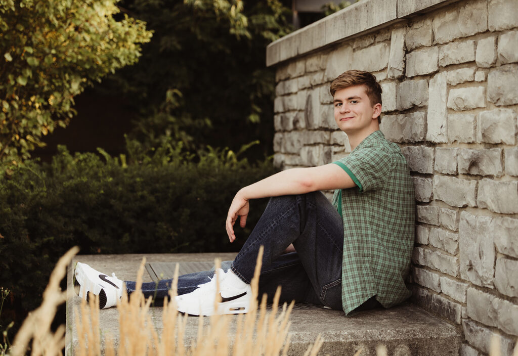 High school senior guy in a green shirt and jeans sitting beside a stone wall during senior pictures