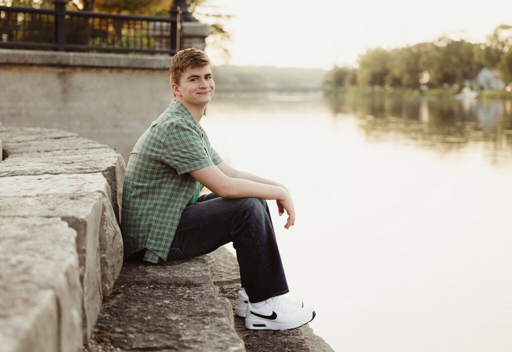 High school senior sitting along the river at sunset during his West Dundee senior photo session