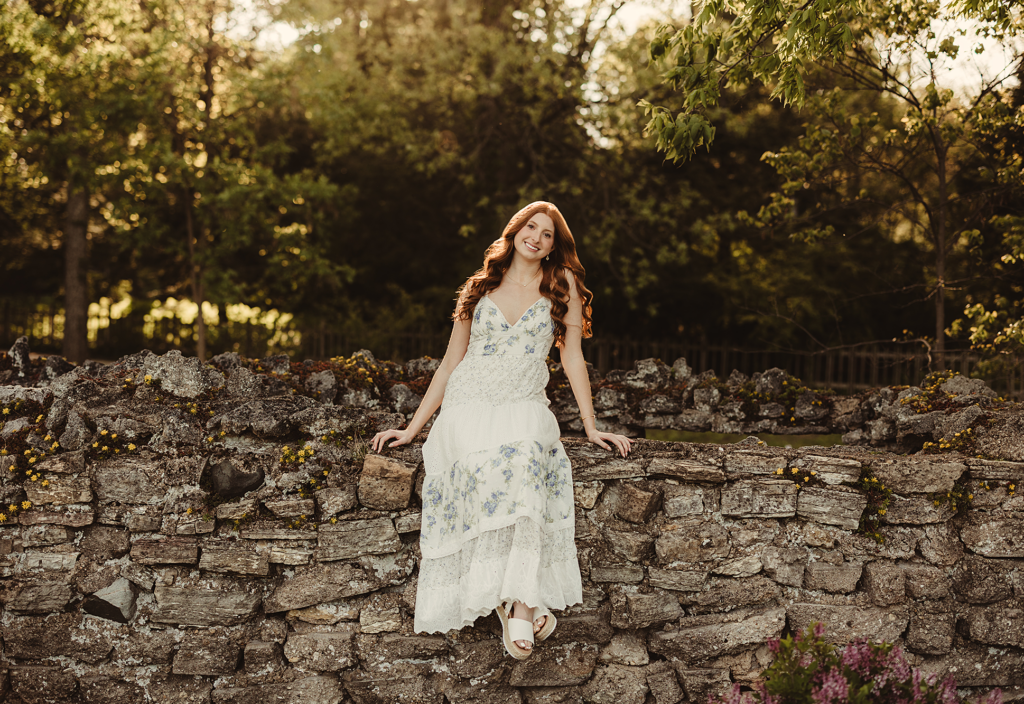 St. Edward Central Catholic High School senior in a white floral dress posing by a stone wall during her senior photo session at Fabyan Forest Preserve
