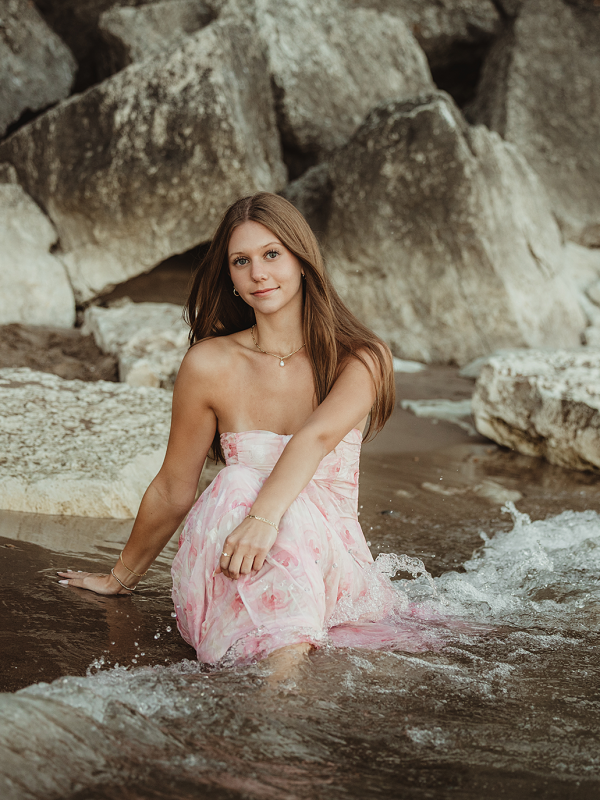 High school senior in pink dress sitting in shallow water at Lighthouse Beach in Evanston, Illinois during sunset photo session