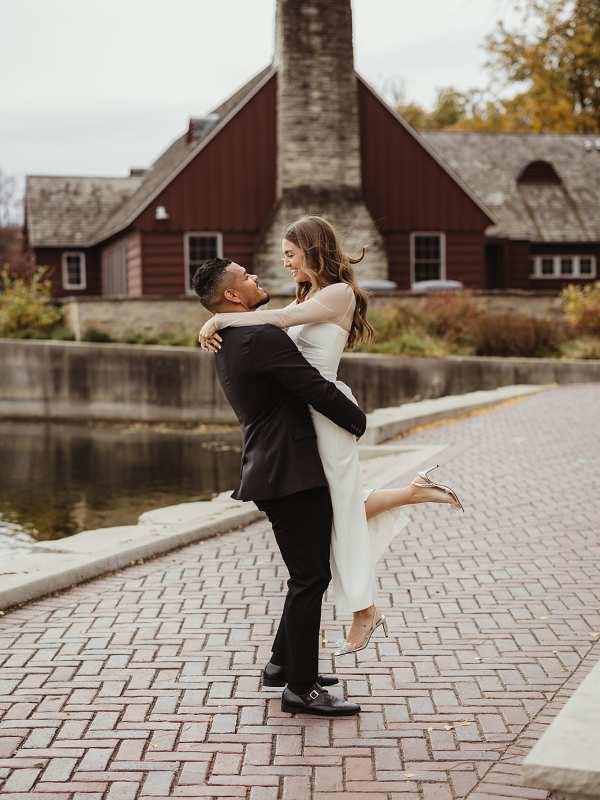 Groom lifts bride along the brick walkway beside the water at Lake Ellyn Boathouse in Glen Ellyn, Illinois during a fall wedding, photographed by LibbyLou Images.
