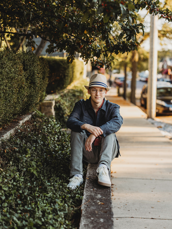 High school senior boy sitting on a stone ledge along a tree-lined sidewalk in downtown St. Charles, Illinois during a golden hour senior photo session by LibbyLou Images.