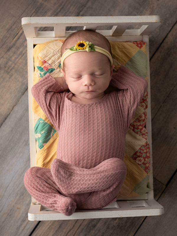 Newborn baby girl wearing a dusty rose outfit while sleeping in a miniature vintage bed